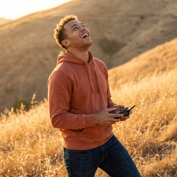 A young man, early-to-mid 20s, light-brown complexion and short curly hair, wearing a rust-orange hoodie and dark jeans, standing on a sun-drenched open hillside at golden hour. He grips the drone controller with both hands, head tilted upward, mouth open in an unguarded laugh of pure delight as the drone ascends out of frame above him. Warm late-afternoon light catches his face and the surrounding dry golden grass. The controller is the only product element visible. Candid, joyful, not staged. Square 1:1, 